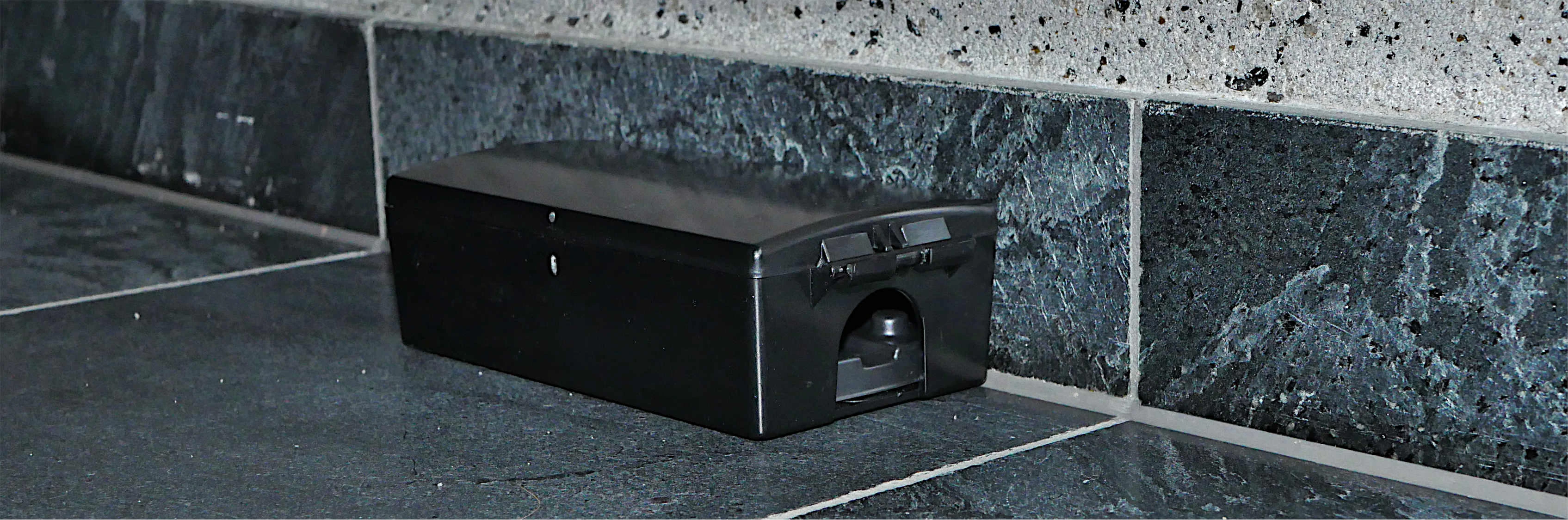 Czarna stacja deratyzacyjna ustawiona przy ścianie na ciemnych płytkach, z widocznym wejściem tunelowym z przodu.