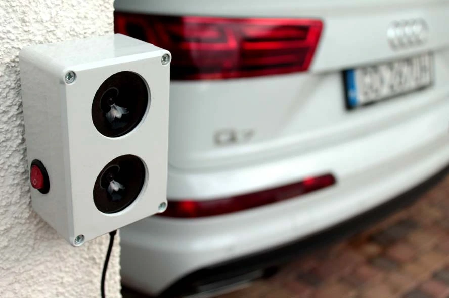 Ultradźwiękowy odstraszacz gryzoni zamontowany na ścianie, w tle tył białego Audi Q7 na podjeździe.