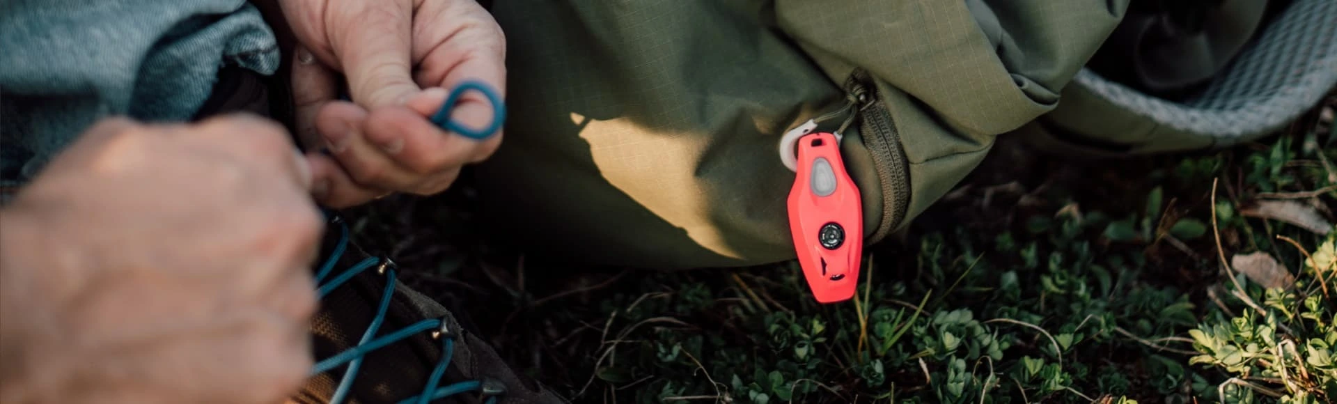 Czerwone urządzenie inMOLESS przypięte do zielonego plecaka, obok osoba wiążąca buty turystyczne w terenie.