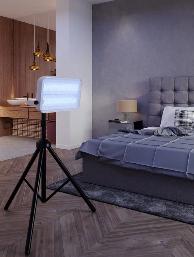 Lampa owadobójcza LED na statywie ustawiona przy łóżku w nowoczesnej sypialni z szarą pościelą i tapicerowanym zagłówkiem.