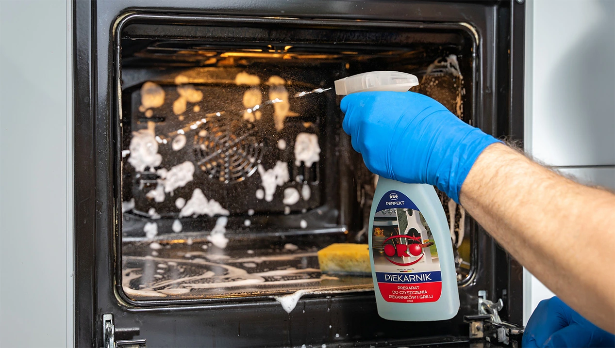 Ręka w niebieskiej rękawiczce rozpyla środek ORO Perfekt do czyszczenia piekarników i grilli wewnątrz zabrudzonego piekarnika.