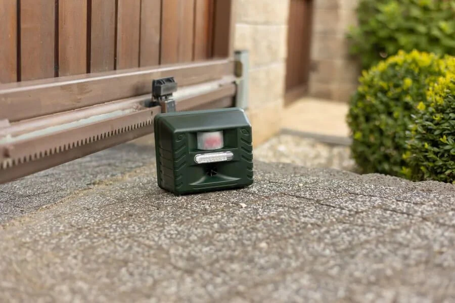 Zielony odstraszacz zwierząt z czujnikiem ruchu i diodą LED, ustawiony na podjeździe przy bramie wjazdowej.