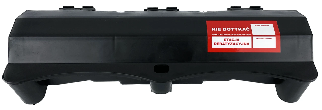 Czarna, podłużna stacja deratyzacyjna z czerwoną etykietą „Nie dotykać – Stacja deratyzacyjna” informującą o obecności trutki na gryzonie.