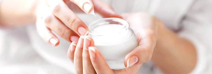 Dłonie kobiety nabierające krem z otwartego, białego słoiczka, w jasnym, miękkim oświetleniu.