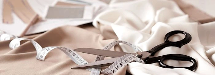 Nożyczki krawieckie i centymetr leżące na beżowym i białym materiale, w tle próbki tkanin i wykroje.