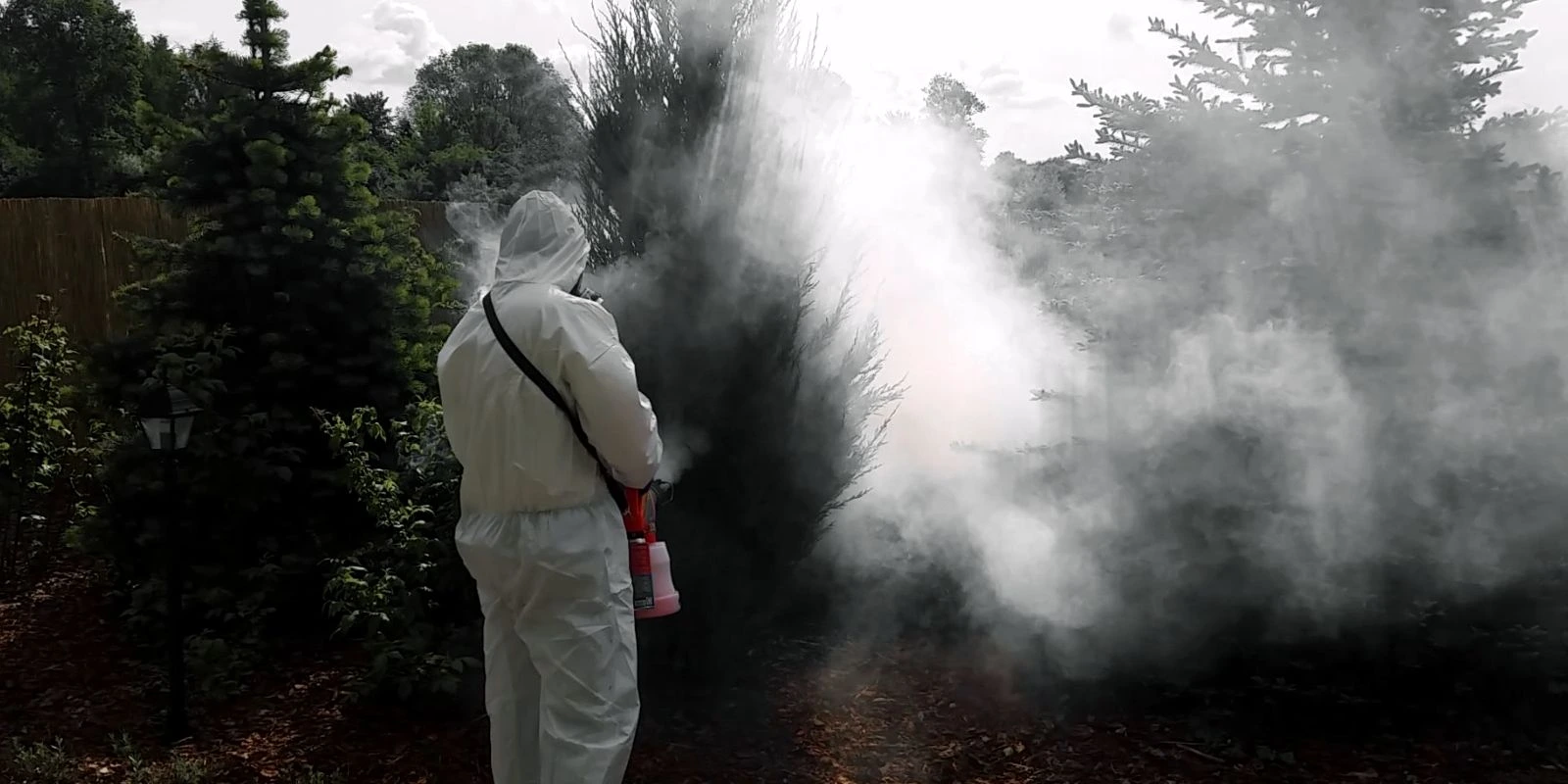 Osoba w kombinezonie ochronnym wykonuje zamgławianie roślin w ogrodzie, otoczona gęstą mgłą opryskową.